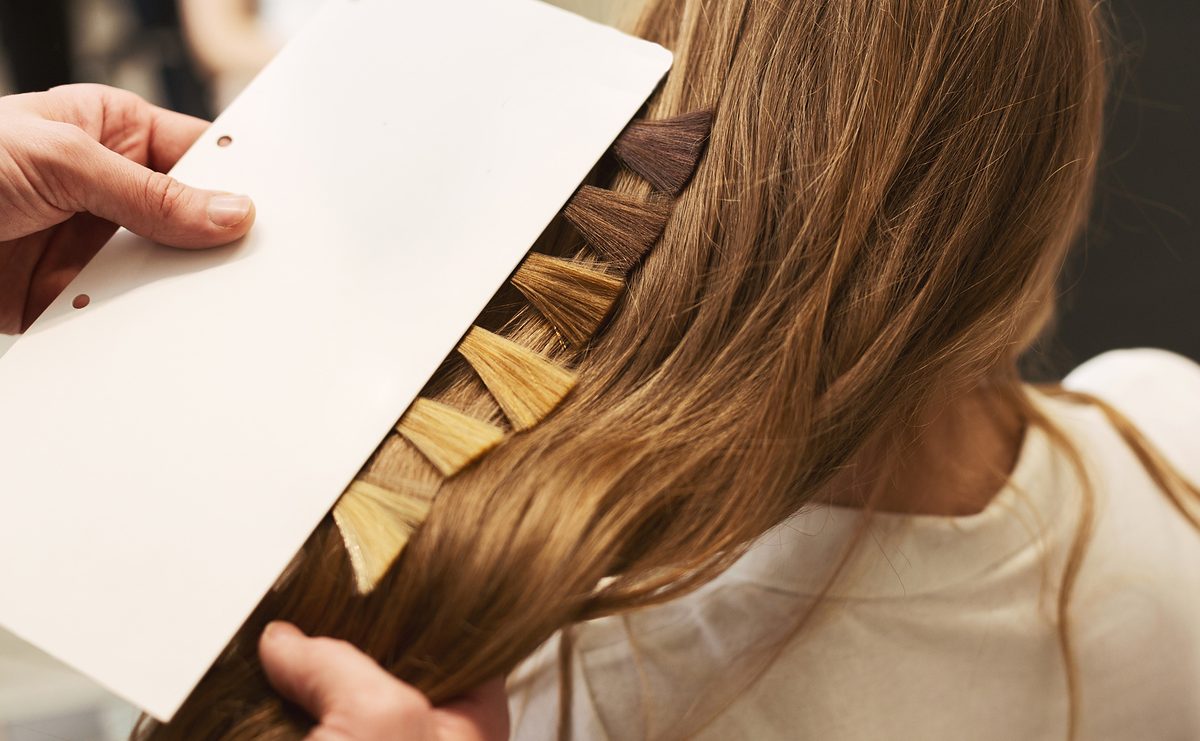 A stylist holds a hair color swatch card beside a client’s long, light-brown hair to compare shades.
