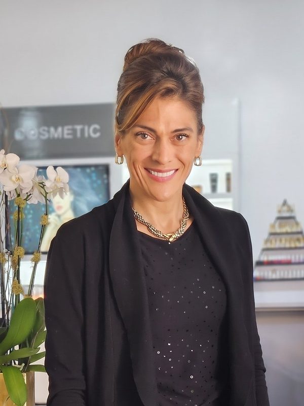 Esther Klein smiling in her salon, wearing black, with orchids and product shelves behind her.