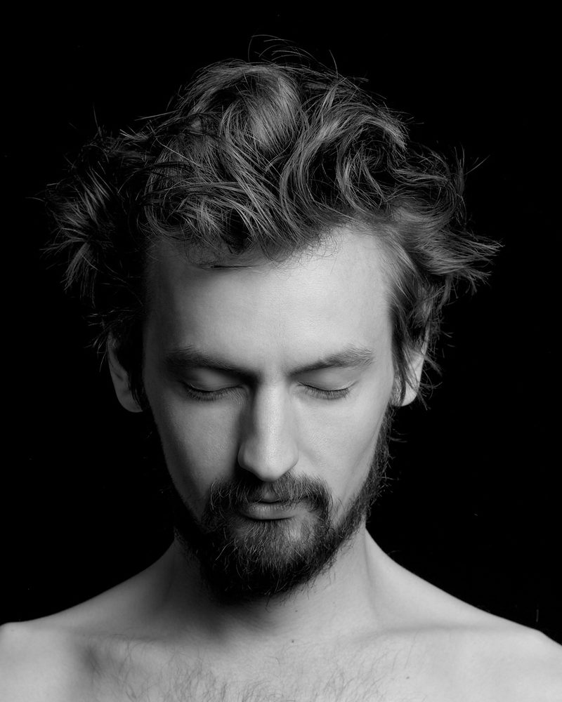 Black-and-white portrait of a bearded man with tousled, textured hair.