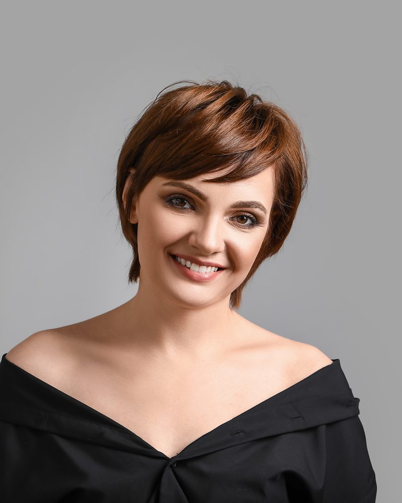 Smiling woman with a short, textured auburn pixie cut and side-swept bangs.
