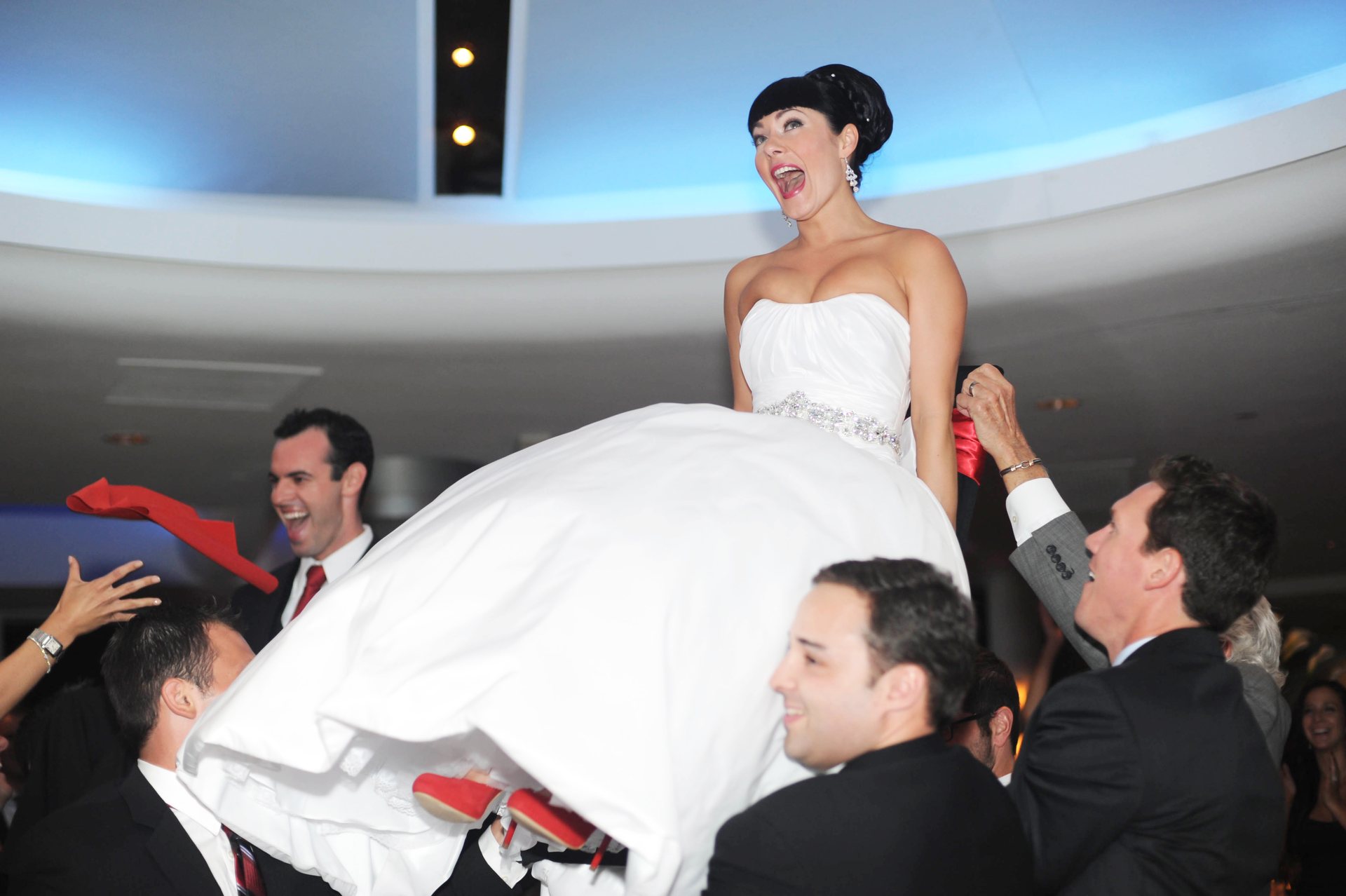 A joyful bride lifted on a chair by her guests during her wedding celebration, her dark hair in a sleek updo with bangs